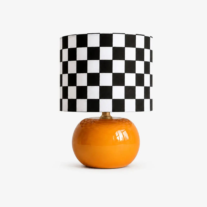 Checkerboard table lamp with glossy orange round base and black and white checkered drum shade, isolated on clean neutral gray background for modern home decor