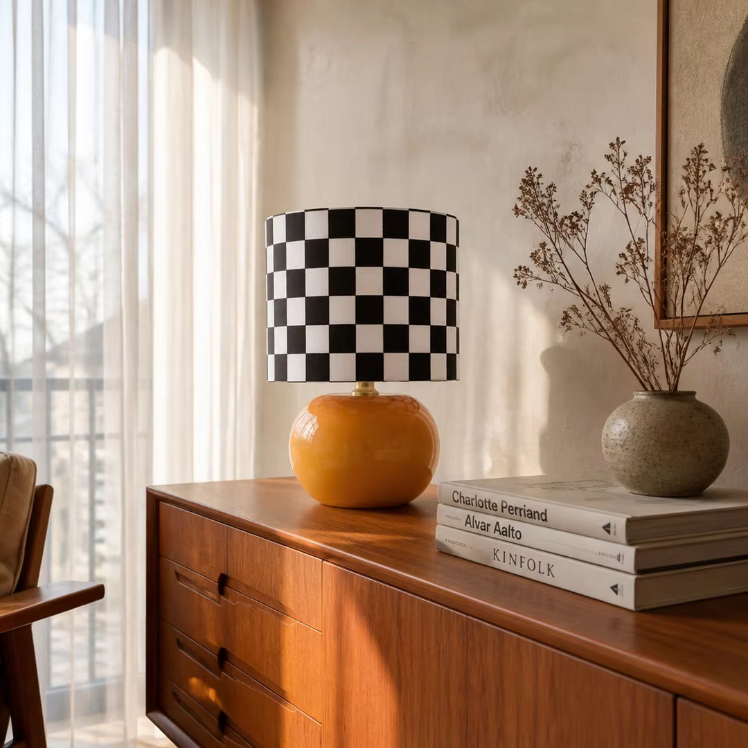 Mid century modern checkerboard table lamp with glossy orange round base and black and white checkered fabric drum shade on a wooden living room credenza