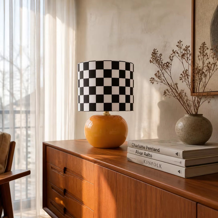 Mid century modern checkerboard table lamp with glossy orange round base and black and white checkered fabric drum shade on a wooden living room credenza