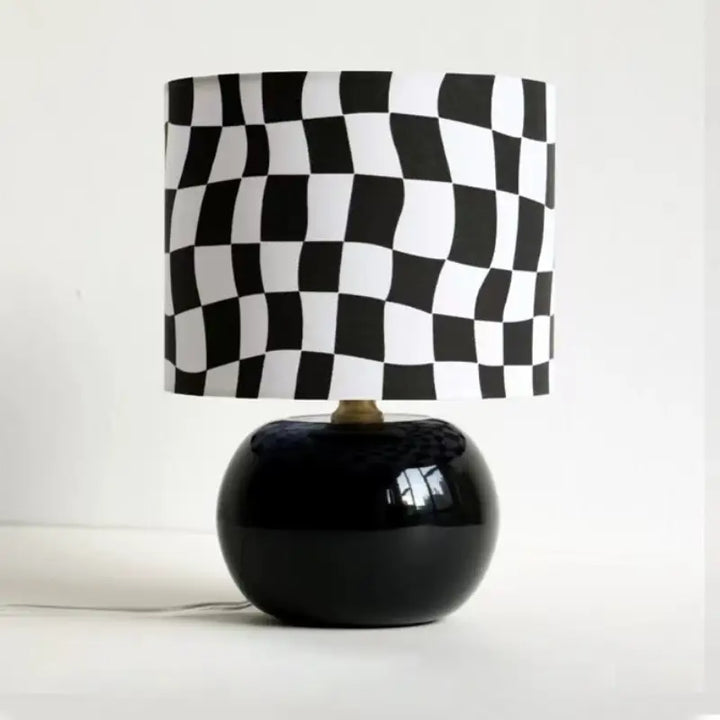 Wavy checkerboard table lamp with glossy black round glass base and psychedelic optical chessboard fabric drum shade on white background