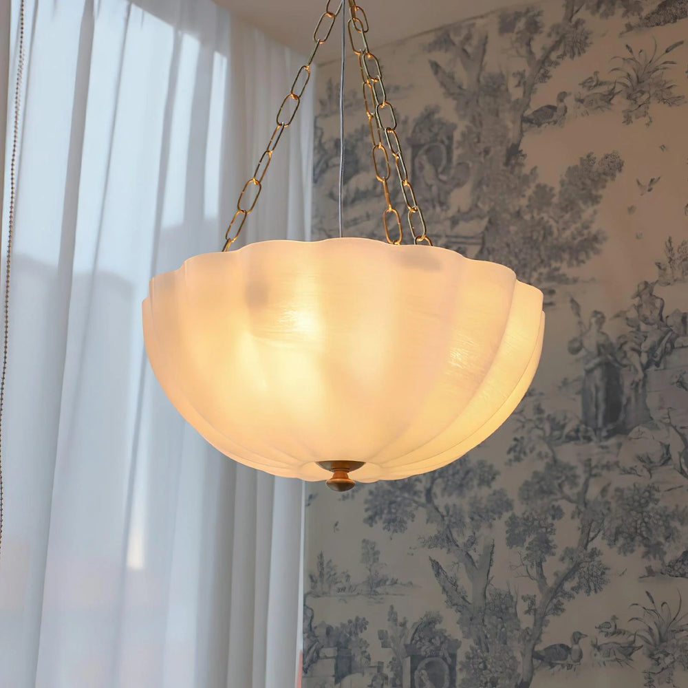Cream frosted glass bowl chandelier with a softly scalloped edge, suspended by gold metal chains, emitting warm ambient light against a backdrop of sheer curtains and patterned wall décor.