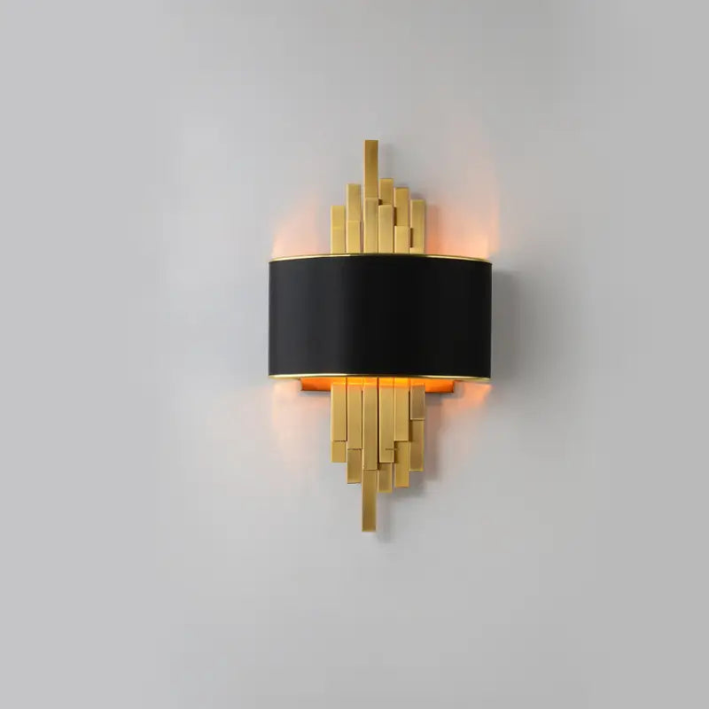 Luxury pipe wall lamp with black fabric drum shade and staggered gold metal strips casting warm ambient light on matte white studio wall