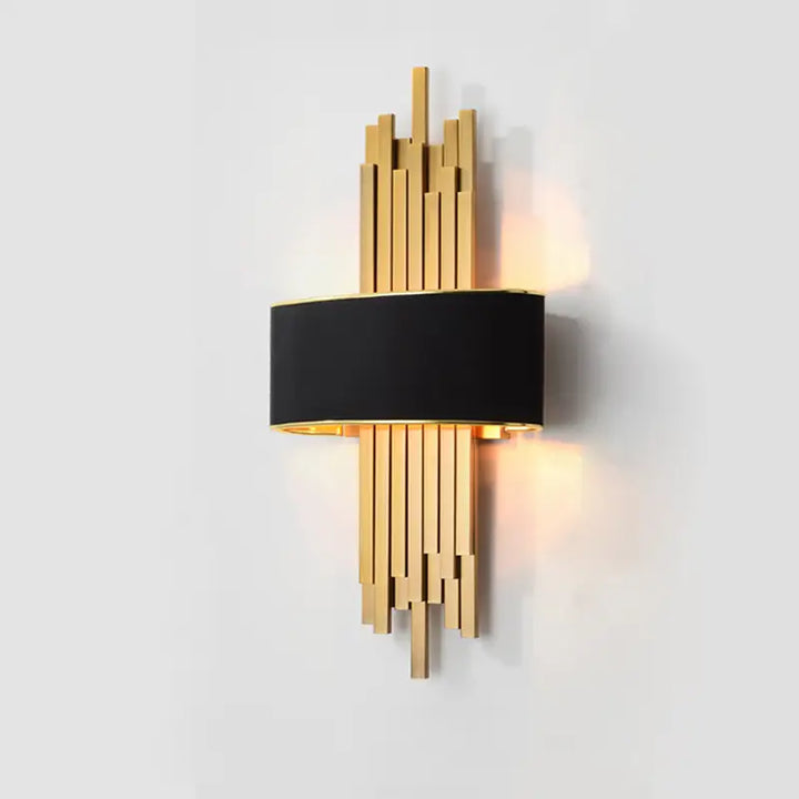 Luxury art deco pipe wall sconce with black fabric drum shade staggered gold metal strips casting soft warm glow on plain light grey wall