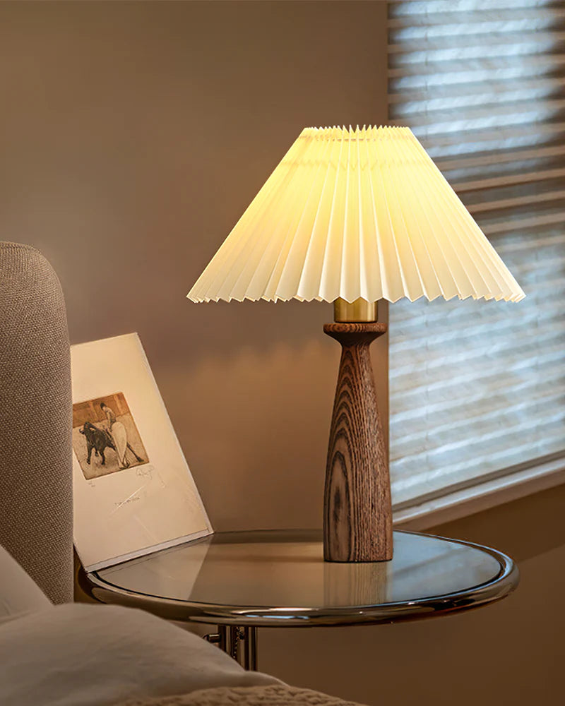 Pleated table lamp turned on with warm soft light, placed on a bedside table creating a cozy nighttime bedroom atmosphere